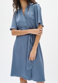 Blaues Wickelkleid aus leichtem, strukturiertem Stoff. Verfügt über kurze Ärmel, einen Bindegürtel in der Taille und einen leicht ausgestellt geschnittenen Saum.