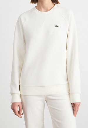 Femme portant un sweat-shirt blanc uni à manches longues et un pantalon blanc assorti, avec un petit logo crocodile vert sur la poitrine gauche.