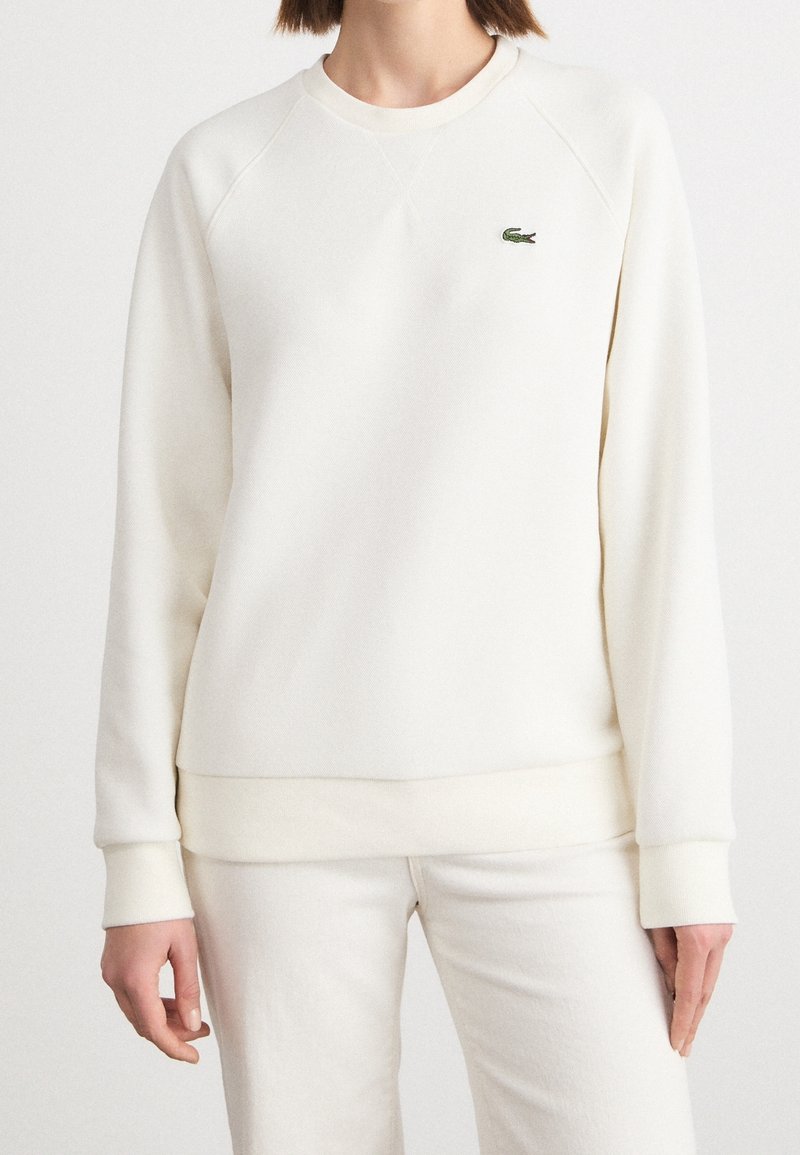 Femme portant un sweat-shirt blanc uni à manches longues et un pantalon blanc assorti, avec un petit logo crocodile vert sur la poitrine gauche.