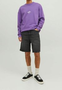 Felpa di cotone viola con logo bianco, abbinata a pantaloni corti in denim neri e sneaker nere. I calzini sono di colore crema e visibili.