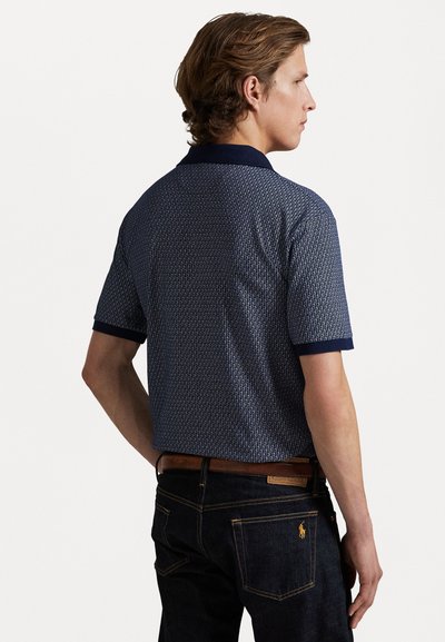 Camisa polo de manga curta azul marinho com um padrão geométrico, colarinho e punhos sólidos. Combinada com jeans escuros e um cinto castanho.