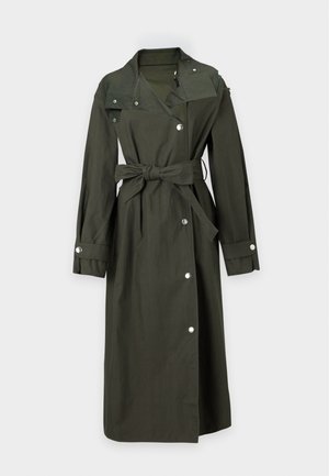 Kvinde iført en lang mørkegrøn trenchcoat med bælte over blå jeans og sorte sko, stående mod en ensfarvet lys baggrund.