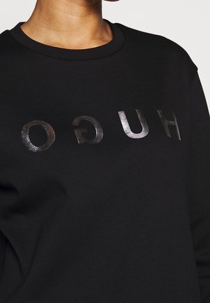 Personne portant un sweat-shirt noir avec un texte brillant et inversé "HUGO" imprimé sur la poitrine.
