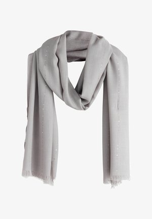 Kvinde iført en gul kjole med blonderkant og et lysegråt sjal bundet om skuldrene, accessorized med et guldarmbånd og øreringe.