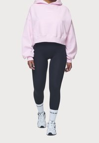 Cropped pink hoodie with a front pocket and white logo, paired with form-fitting black leggings and white athletic socks, worn with sneakers.