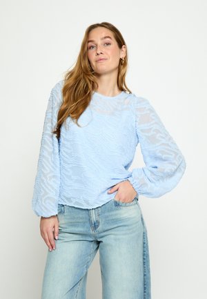 Kobieta z długimi włosami, ubrana w jasnoniebieską teksturowaną bluzkę i jasnoniebieskie dżinsy, stojąca z jedną ręką w kieszeni na białym tle.