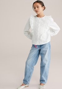 Valge pluus tekstuursete täpikestega, volangiliste õlgade ja pitsdetailidega, kombineeritud helesiniste laia säärega teksapükstega ja sädelevate kingadega.