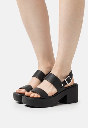 Black platform sandals with wide leather straps and a buckle accent. The sole is thick and textured for grip and support.
