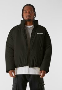 Sort pufferjakke med høj krave, lynlås lukning og et logo patch på venstre bryst. Bæres over en grå sweater og en hvid skjorte.
