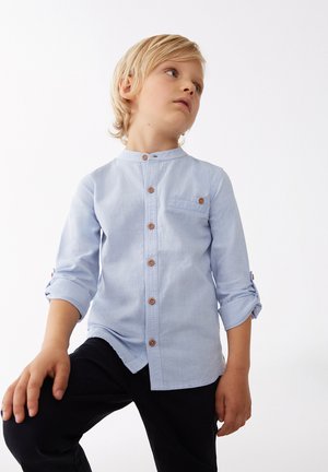 Camisa de botones de manga larga azul claro con cuello alzado, bolsillo en el pecho, botones marrones y puños de manga enrollados. Llevada con pantalones oscuros.
