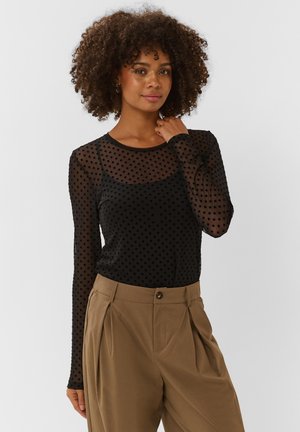 Mujer con cabello rizado lleva una blusa negra transparente de manga larga con lunares y pantalones marrones sueltos de cintura alta sobre un fondo liso.