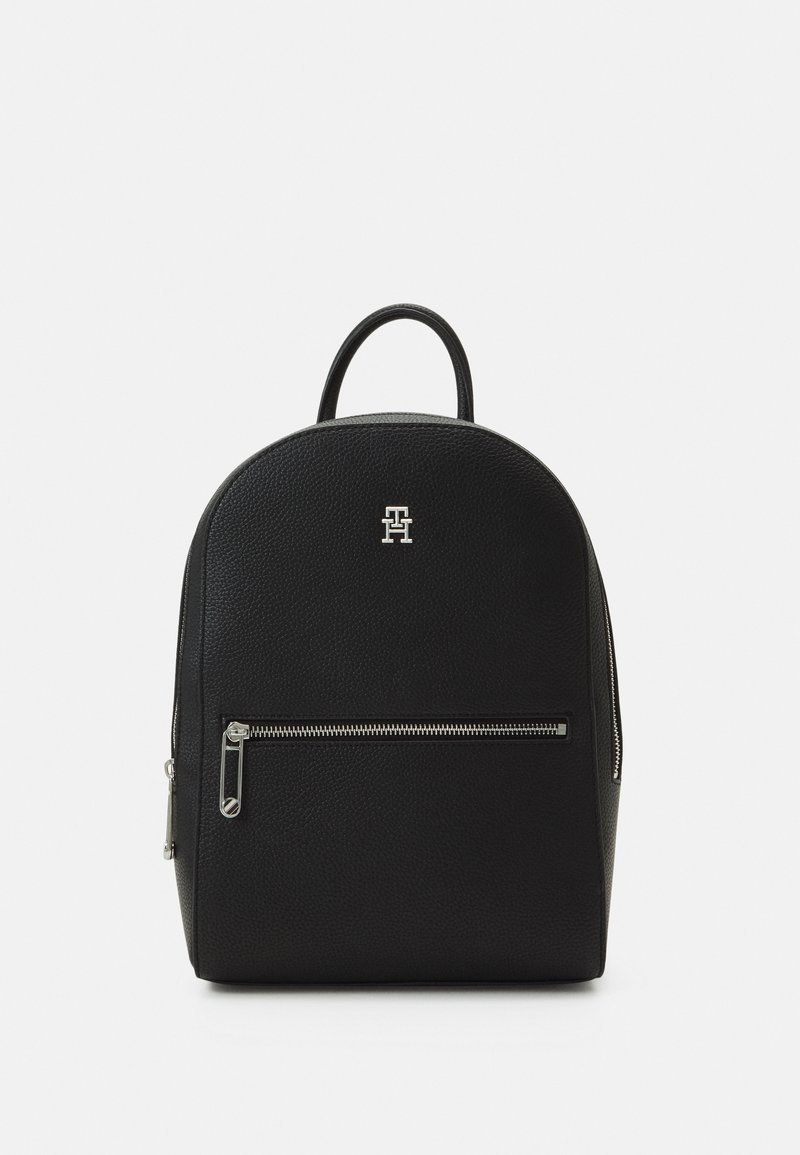 Sac à dos noir texturé avec poche à fermeture éclair argentée sur le devant et petit logo argenté "TH" au-dessus de la poche, présenté sur fond blanc.