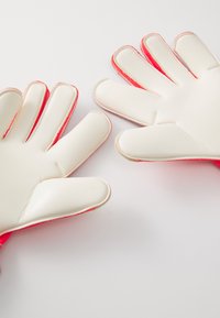 Et par hvite og røde keeperhansker lagt flatt på en hvit overflate med fingrene spredt ut.