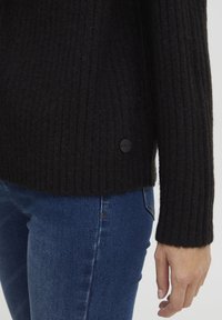 Maglione nero a coste realizzato in materiale a maglia testurizzato, con un dettaglio di bottone nero rotondo sull'orlo e maniche lunghe. Indossato con jeans blu.