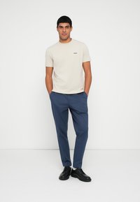 T-shirt bege de mangas curtas com um logotipo preto, combinada com calças sob medida azul-marinho e sapatos pretos de atacadores, posando contra um fundo branco simples.