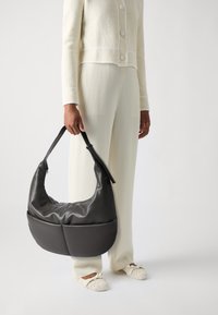Sac hobo en cuir noir au design incurvé, avec une large anse et un discret détail de fermeture éclair. Porté avec des vêtements texturés de couleur claire.