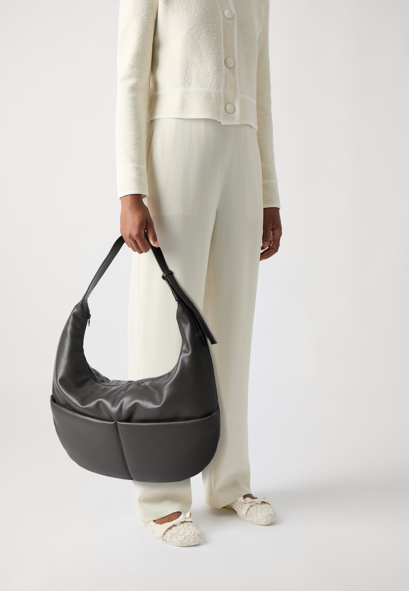 Sac hobo en cuir noir au design incurvé, avec une large anse et un discret détail de fermeture éclair. Porté avec des vêtements texturés de couleur claire.