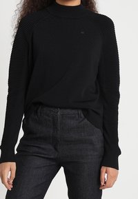 Svart ribbad tröja med hög krage och långa ärmar, i kombination med mörka jeans. Texturerad tyg på axlarna ger detalj.