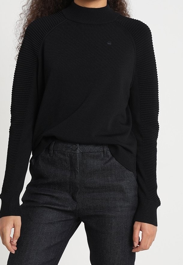 Svart ribbad tröja med hög krage och långa ärmar, i kombination med mörka jeans. Texturerad tyg på axlarna ger detalj.