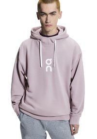 Svetloružová mikina s kapucňou a predným logom, vybavená sťahovacou šnúrkou a dlhými rukávmi, skombinovaná so sivými nohavicami. Hladká textúra a voľný strih.