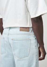 Osoba o ciemnej karnacji ubrana w jasnoniebieskie dżinsy z metką "EIGHTYFIVE" oraz luźną białą koszulkę z krótkim rękawem, widziana od talii w dół.