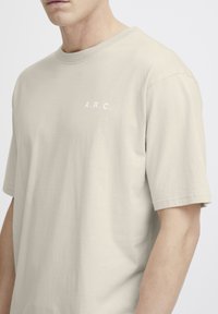 Beige bomull t-skjorte med rund hals, korte ermer og hvit "A.R.C." tekst på venstre bryst. Glatt tekstur, klassisk passform.