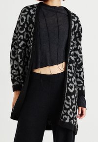 Cardigan de leopardo en negro y gris, diseño abierto, con textura de punto, de mangas largas y puños acanalados, superpuesto a un top negro corto y pantalones.