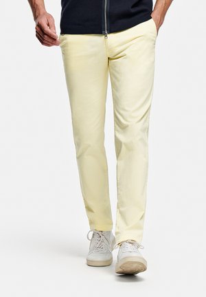 Man loopt en draagt lichtgele slim-fit broek, witte sneakers en een donker jack met ritssluiting, handen gedeeltelijk in de zakken.