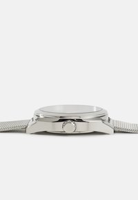 Reloj de acero inoxidable plateado con una caja redonda lisa, cara de cristal y una pulsera de malla metálica trenzada, con una corona en el lateral.