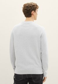 Maglione grigio chiaro con texture, maniche lunghe e scollatura rotonda, con polsini e fondo a costine. Indossato sopra pantaloni scuri.