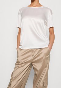 Satinowa biała bluzka z krótkim rękawem i okrągłym dekoltem w zestawie z błyszczącymi, jasnobeżowymi spodniami o szerokich nogawkach i kieszeniami. Gładka faktura na całej powierzchni.
