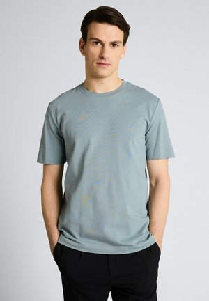 Młody mężczyzna stojący z rękami w kieszeniach, ubrany w jasnoniebieską koszulkę z krótkim rękawem i czarne spodnie na jednolitym tle.