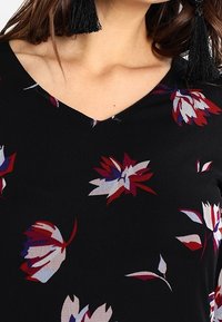 Svart tygtröja med V-ringning och djärva blommönster i röd, vit och blå. Inkluderar framträdande svarta fransdetaljer.