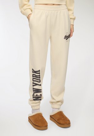 Pantaloni della tuta crema con vita elasticizzata, caratterizzati da stampe in marrone scuro con la scritta "Brooklyn" e "NEW YORK". Accompagnati da pantofole marroni senza tacco.