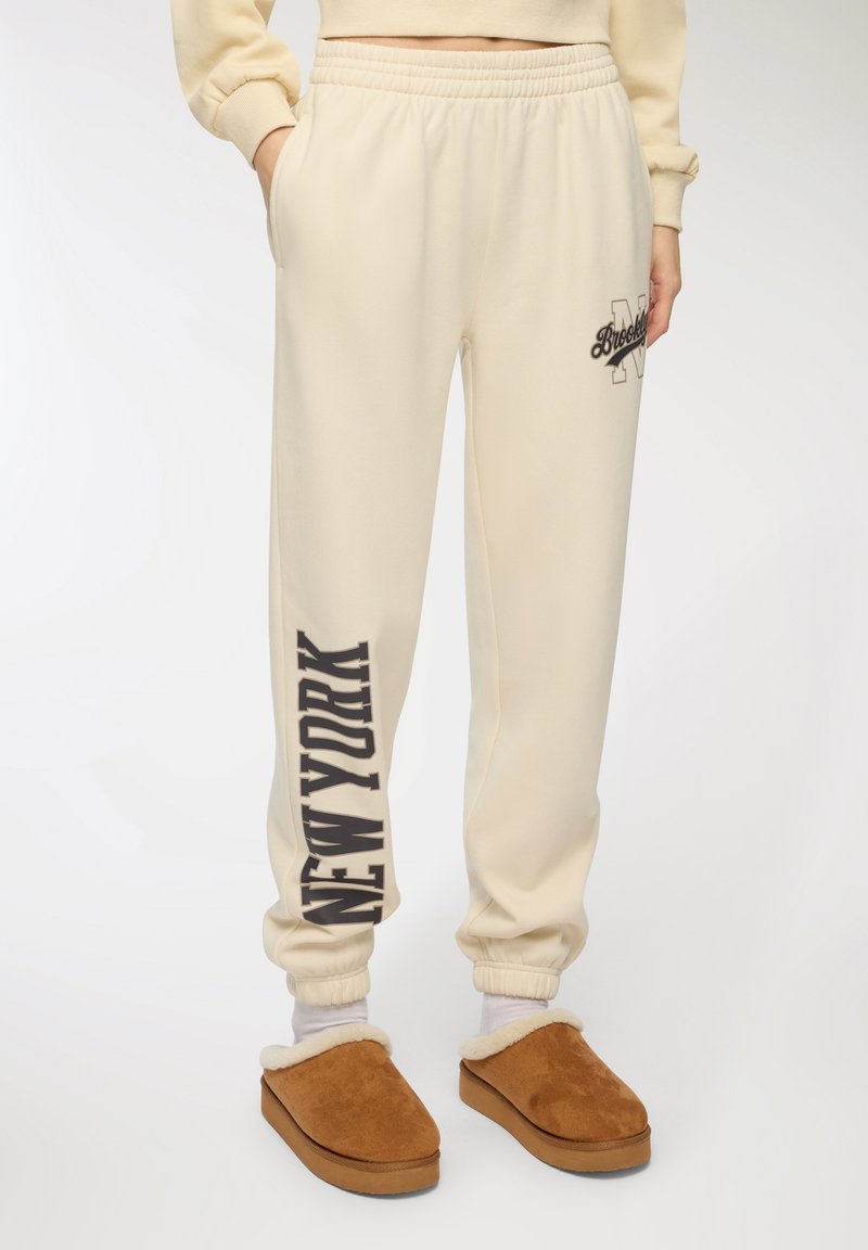 Kremowe spodnie dresowe z elastycznym pasem, z nadrukami "Brooklyn" i "NEW YORK" w ciemnobrązowym kolorze. W komplecie brązowe klapki.