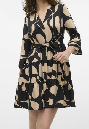 Femme portant une robe à motif abstrait noir et beige, avec des manches trois-quarts et une ceinture nouée à la taille, debout devant un fond uni.