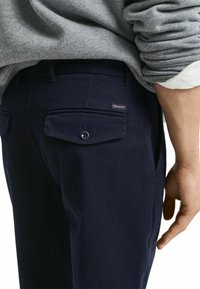 Pantalons en coton bleu marine avec une poche arrière à bouton, présentant une petite étiquette logo. La texture est lisse avec une coupe classique.