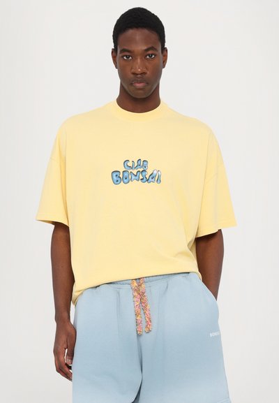 Geel katoenen T-shirt met een ronde halslijn en korte mouwen, voorzien van een blauwe grafische tekst "CLAB BONSAI" op de voorkant. Lichtblauwe shorts met een gestructureerde koord.