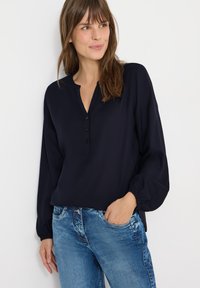 Blouse bleu marine à manches longues avec un col en V et une patte de boutonnage à quatre boutons, caractérisée par une coupe décontractée et des poignets élastiqués, associée à un jean bleu clair.