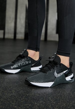 Persona che indossa scarpe da allenamento Nike Metcon nere e bianche e leggings compressivi neri in piedi sul pavimento di una palestra.