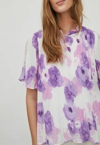 Veckad blommig blus i vitt, med stora lila och rosa blommor. Lättviktig tyg med korta ärmar och rund halsringning.