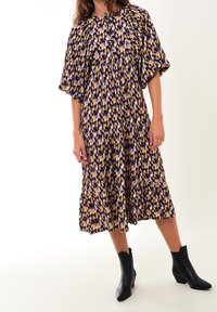 Vestido de patrón púrpura, amarillo y rosa con un corte holgado, mangas cortas abullonadas, diseño en capas y largo hasta la rodilla. Usado con botas negras hasta el tobillo.