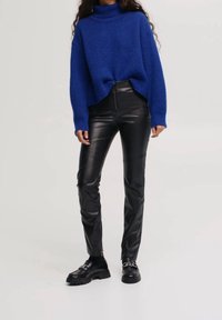 Pull à col roulé tricoté bleu à coupe ample, associé à un pantalon skinny en similicuir noir. Chaussures épaisses noires avec un détail en chaîne.
