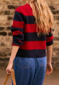 Suéter a rayas rojas y marinas con cuello redondo y mangas enrolladas, combinado con jeans de pierna ancha de color azul claro y una bandolera beige.