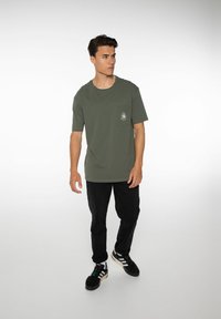 Olijfgroene katoenen t-shirt met een ronde hals en korte mouwen, voorzien van een borstzakje en een klein logo in het wit. Draagt met zwarte broek en schoenen.