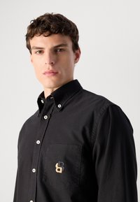 Joven con cabello corto y rizado que lleva una camisa negra de botones con un logo bordado en beige y negro en el bolsillo del pecho.