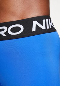 Detailní záběr na pas s modrými sportovními kraťasy Nike Pro a černým pasem s bílým nápisem "NIKE".