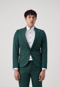 Costume vert foncé avec une veste sur mesure et un pantalon assorti, associé à une chemise blanche à boutons noirs. Texture de tissu lisse et soyeuse.