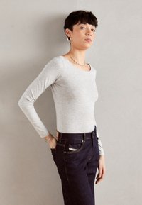 Grauer, langärmeliger, figurbetonter Oberteil aus weichem Material, kombiniert mit dunklen Denim-Jeans mit Gesäßtaschen und einem Etikett am Bund.