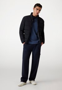 Zwarte pufferjack met hoge kraag, blauwe longsleeve shirt eronder, marineblauwe cargo pants met ritsen, en witte sneakers.